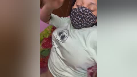Media: Video of a person in a light green shirt with a cartoon character logo, wearing a black and white patterned face mask, taking a selfie. Background includes colorful quilted fabric and blurred indoor setting.