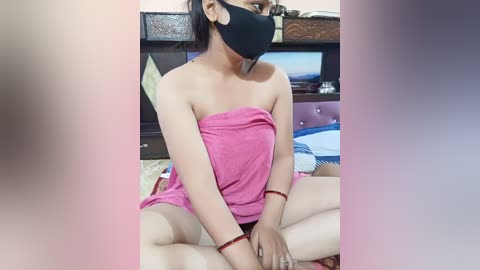 Video of a woman with light skin and dark hair wearing a black face mask, pink strapless dress, and red bracelets, sitting cross-legged on a bed with a wooden headboard and TV in the background.