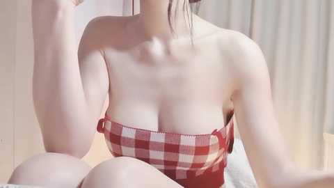 Video of an Asian woman with fair skin, wearing a red and white checkered strapless top, sitting on a bed with white sheets. The background features a blurred, soft-focus setting.