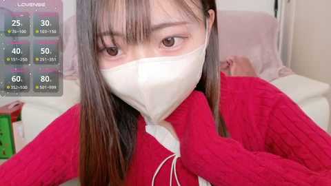 Video of an East Asian woman with long black hair, wearing a red knitted sweater and a white face mask, lying in bed. Background includes a pink pillow and a digital temperature display.