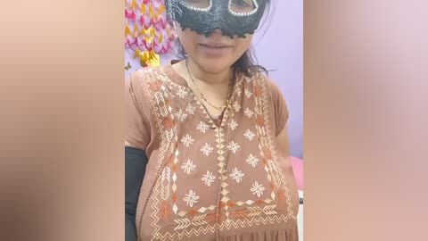 Video of a woman with medium skin tone, wearing a black lace mask, beige embroidered top, and a black long-sleeved shirt. Background features colorful paper flowers and a pink wall.