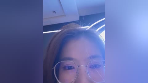 Video of a young Asian woman with straight brown hair, wearing round glasses, taken from a low angle, highlighting her face. She is indoors, with a ceiling light and dark walls in the background.