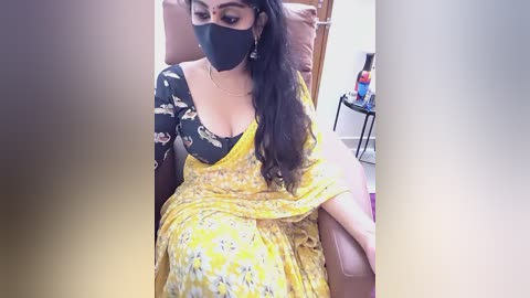 Video of a South Asian woman with medium skin tone, long dark hair, and a black face mask, wearing a yellow floral saree with a black floral blouse, seated in a pink leather chair in a room with a white wall and a wooden door.