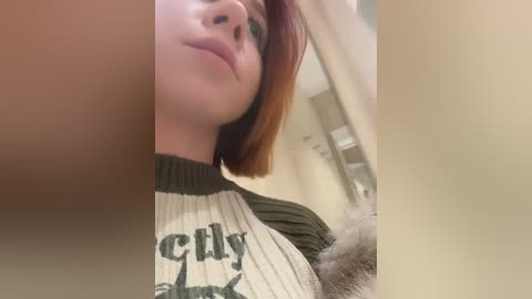 A close-up video of a young woman with straight, shoulder-length auburn hair, wearing a ribbed, green and cream top with \"Gently\" printed on it. The background is blurry, showing a beige wall and a bathroom with a white towel.