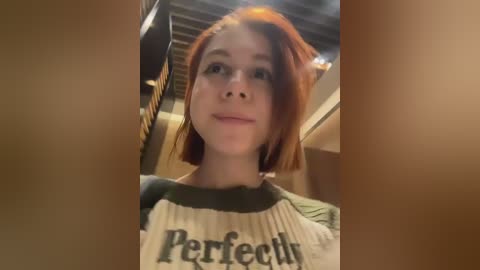 Video of a young woman with short red hair and pale skin, wearing a \"Perfectly\" t-shirt, standing in a dimly lit room with exposed ceiling beams.
