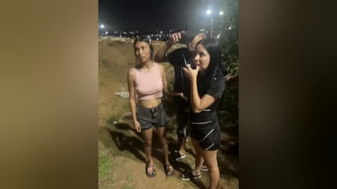 A video of two women in a dark, urban street at night. One woman in a pink crop top and denim shorts, the other in black athletic gear, taking a selfie.