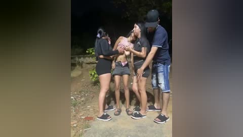 Video of a group of four people, two women and two men, engaged in a playful embrace on a dark, outdoor path, wearing casual attire.