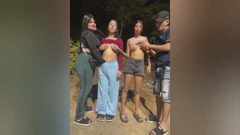 Video of four women in a dark outdoor setting, all topless, with one holding another's breasts. They wear casual attire, including high-waisted pants and sneakers, standing closely together.