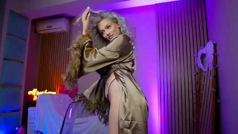 Video of a slender, light-skinned woman with curly platinum blonde hair in a silky, nude satin robe, striking a pose against a purple-lit wall with a white fan, in a modern, dimly-lit room.