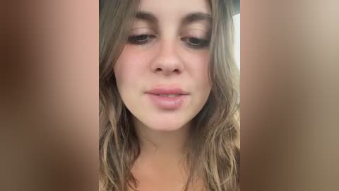 Close-up video of a young woman with light skin, wavy brown hair, and subtle makeup, gazing straight ahead, surrounded by a blurred, warm-toned background.