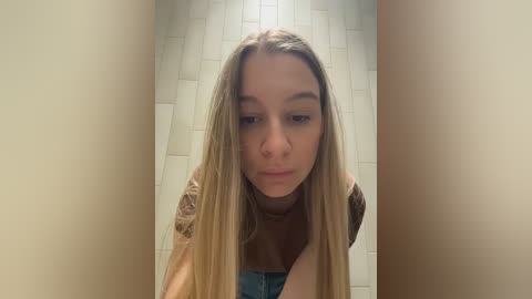 Video of a young woman with long, straight blonde hair, wearing a brown turtleneck and blue jeans, looking serious, in a narrow, tiled hallway with beige walls.