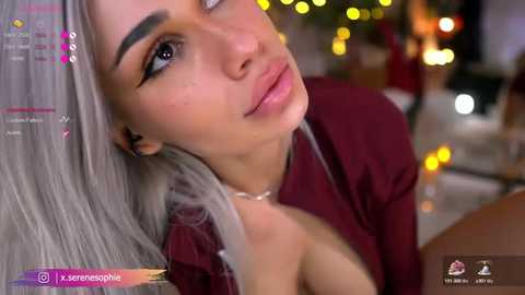 Video of a young woman with fair skin, long platinum blonde hair, and full lips, wearing a burgundy top, in a dimly lit room with blurred, warm-colored background lights.