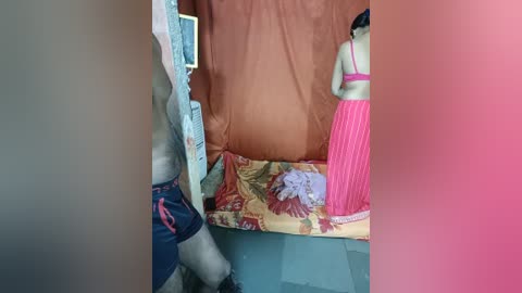 Video of a small, cluttered room with a pink and blue striped wall, featuring a doll wearing a pink dress and a floral shirt on a bed with a floral duvet.