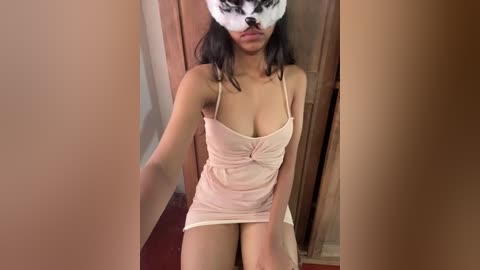 Video of a young Asian woman with medium skin tone and dark hair, wearing a beige mini dress and a white mask with black spots, sitting on a wooden floor, indoors.