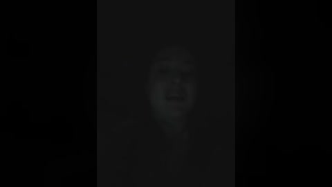 A dimly lit, grainy video of a young man with dark hair and a solemn expression, standing in front of a black background, creating a mysterious, ominous atmosphere.