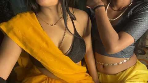 Video of two South Asian women, one with a yellow sari and black bra, the other in a gray crop top, both wearing necklaces, indoors.