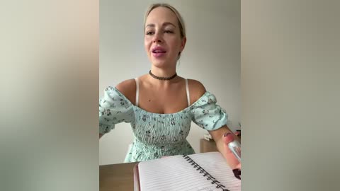 Video of a fair-skinned, blonde woman with a light blue floral dress, off-shoulder, holding a spiral notebook, seated indoors against a plain beige wall, smiling.