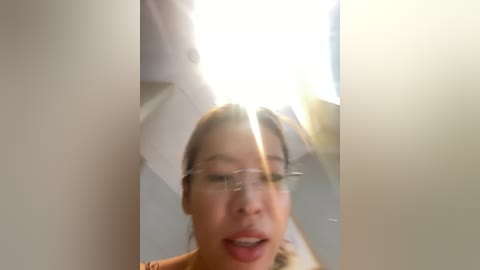 Video of a young Asian woman with light skin and glasses, smiling broadly with parted lips, captured from a low angle. She is indoors, with a bright, sunlit ceiling and a soft focus background.