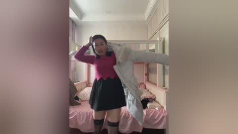 Video of an Asian woman adjusting her white jacket in a pastel-colored bedroom with a pink bedspread and floral patterns.
