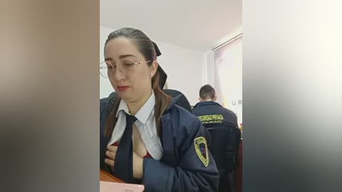 Video of a young woman with glasses, in a dark navy school uniform with a white shirt, sitting in a classroom. Two police officers are present in the background, one with the word \"POLICIA\" on their jacket.