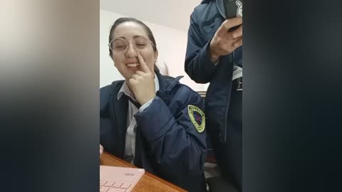 Video of a female police officer with dark hair and glasses, seated at a desk, making a funny face. She wears a navy blue uniform with a badge. Another officer is partially visible in the background.