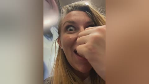 Video of a blonde woman with light skin, wearing a gray top, covering her mouth with her right hand, looking shocked. Background is blurry with an indistinct blue and white pattern.