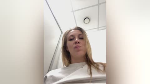 Video of a blonde woman with fair skin and straight hair, wearing a white shirt, standing in a white-tiled bathroom with fluorescent lights, giving a low-angle, upward-looking shot.