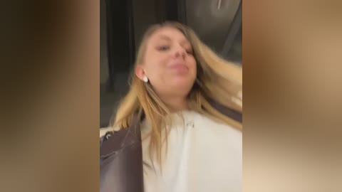 A blurry video of a blonde woman in a white shirt, leaning against a wall with her hair tousled, hands behind her head.