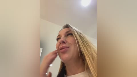 Media: Video of a Caucasian woman with long, straight blonde hair, wearing a white shirt, smiling and playfully touching her nose. Background shows a plain, white-walled room with a ceiling light.