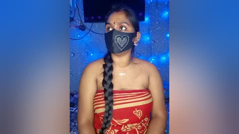 Video of an Indian woman with medium brown skin, wearing a black face mask with a heart design, a red saree with gold embroidery, and long braided hair. She stands in a dimly lit room with blue LED lights on the walls.