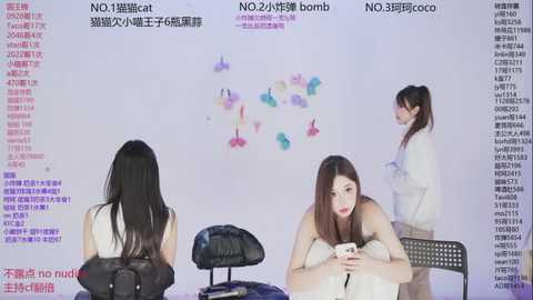 Video of three young Asian women in casual white tops, seated on black chairs, against a white backdrop with colorful balloons, Chinese text, and an empty table.