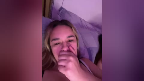 A video shows a young woman with blonde hair, light skin, and a slight smile, holding her nose, lying in a bed with purple pillows.