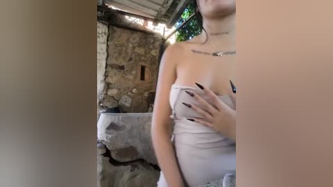A video shows a pregnant woman with a light complexion, wearing a strapless, light-colored dress, standing in a rustic, stone-walled room. She has long, dark hair, and her hands cover her chest.