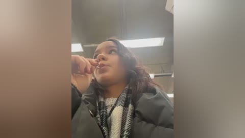 Video of a young woman with light skin and dark hair, wearing a gray jacket and white scarf, seated in a dimly lit train car.