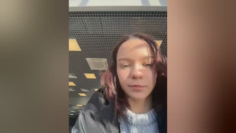 A video of a young woman with shoulder-length auburn hair, wearing a black jacket and a light gray sweater, indoors under fluorescent lighting, her face partially shadowed.