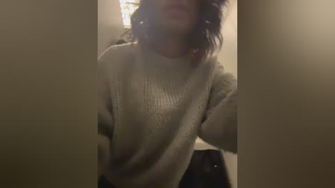 Video of a person with shoulder-length curly hair, wearing a cream-colored, knitted sweater, partially obscured by a dark object in the foreground. The background is dimly lit, with a beige wall.