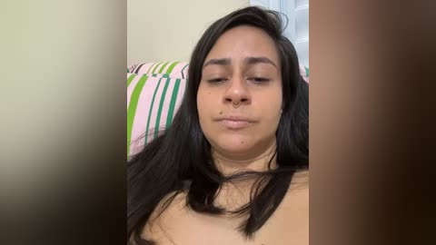 Video of a young woman with medium-brown skin and long, straight black hair, wearing a nose ring, lying on a bed with striped pillows.