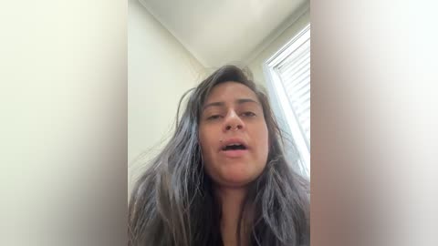 A video of a young woman with long, messy gray hair, light skin, and a slightly open mouth, captured from a low angle. She stands in a dimly lit room with cream-colored walls and a window with white blinds.
