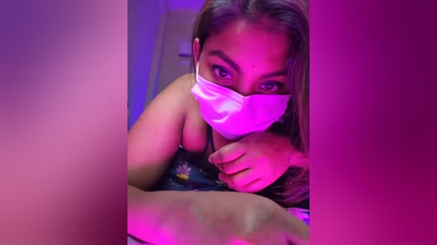 A close-up video of a woman with medium brown skin and long black hair, wearing a white surgical mask and a dark top, against a purple-lit background.