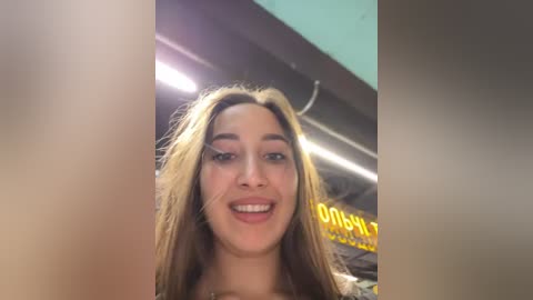 A video of a smiling young woman with long, straight brown hair and fair skin, taken indoors near a brightly lit sign.