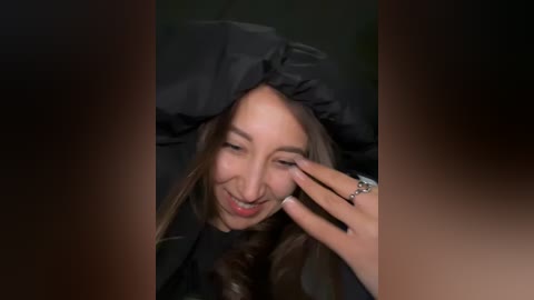 Video of a smiling Asian woman with long brown hair, wearing a black hat, touching her eye with her left hand, against a dark, blurred background.