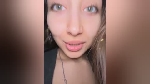 A close-up video of a woman with light brown skin and green eyes, wearing a black top and a silver necklace, captured in a blurry, intimate indoor setting.