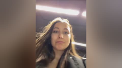 Video of a young woman with long, wavy brown hair, wearing a dark jacket, captured from a low angle, with a blurred background featuring fluorescent lighting.