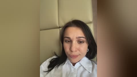 A video of a young woman with light skin, dark hair, and brown eyes, wearing a white shirt, seated in a padded beige chair.