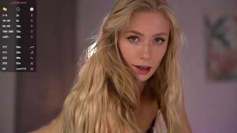 Video of a young, blonde woman with wavy hair, fair skin, and light makeup. She's indoors, wearing a sleeveless top, with blurred background.