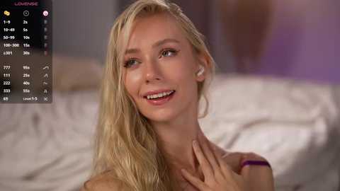 Video of a smiling, nude blonde woman with fair skin, medium build, and small breasts, wearing only a purple bracelet, in a dimly-lit bedroom with blurred background.