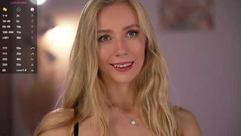 Video of a smiling, fair-skinned blonde woman with long hair, wearing a black top, against a blurred, softly lit background.