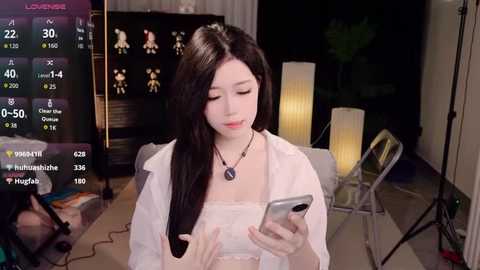 Video of an Asian woman with long black hair, wearing a white shirt, holding a smartphone, in a dimly lit room with a monitor displaying social media metrics.