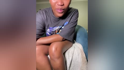 A video of a dark-skinned woman with medium-sized breasts, wearing a gray graphic t-shirt, sitting with knees drawn up, her face partially obscured by her arms.