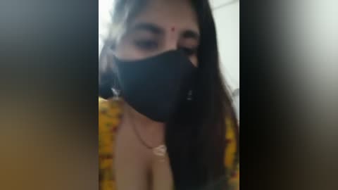 Video of a woman with long dark hair, wearing a black mask and a yellow patterned shirt, partially covered by her hair.